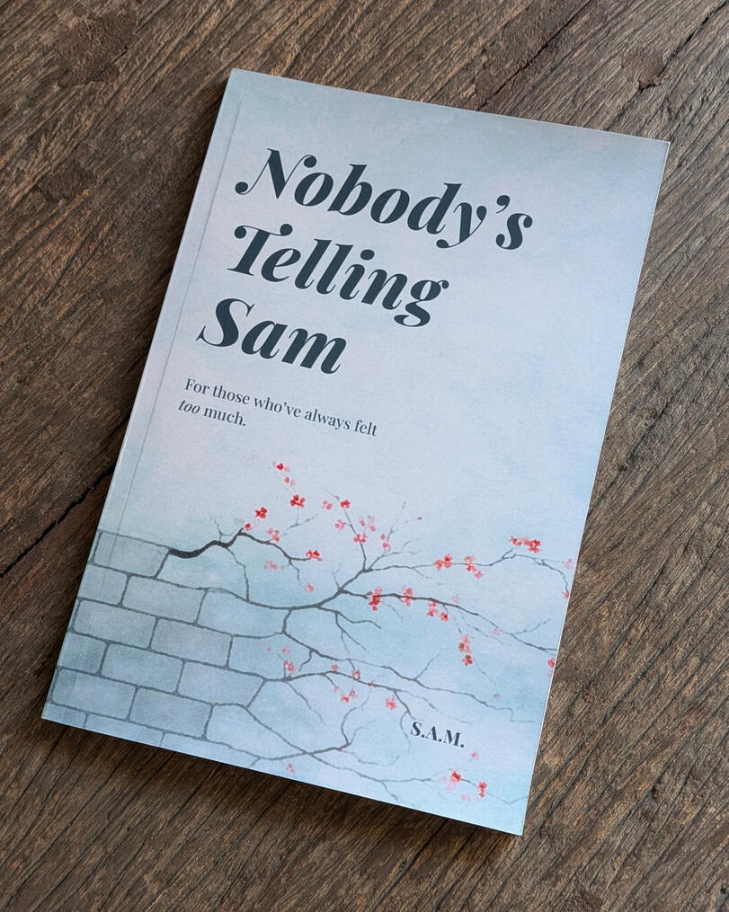 Something real to hold onto. The front cover of 'Nobody’s Telling Sam' resting on a rustic wooden surface, highlighting the book's narrow binding and the natural grain of the wood.